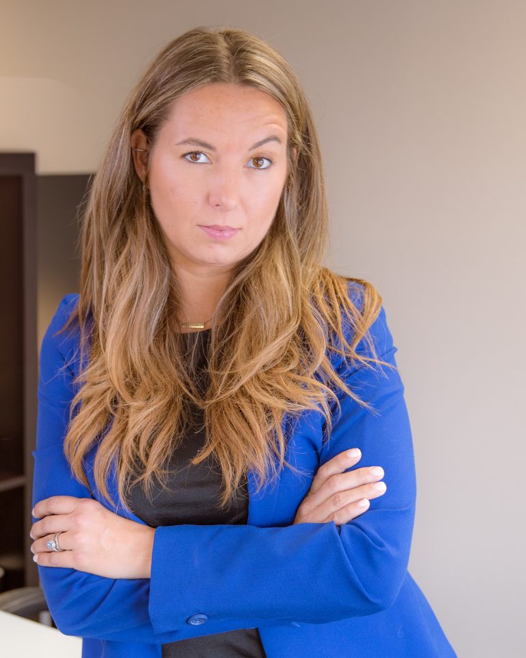 Woman with long hair wearing a blue blazer and black top, standing with arms crossed.
