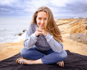 Woman with long hair sitting cross-legged on a black blanket at the edge of a cliff overlooking the ocean.
