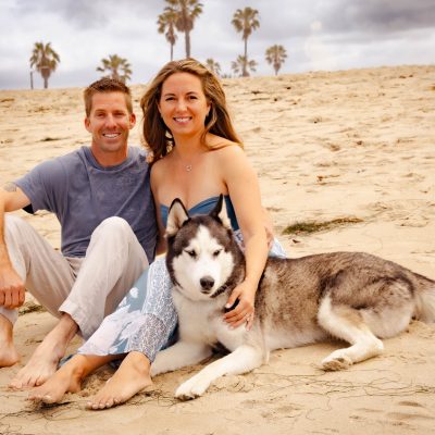 A man and woman sitting on the sand with a Siberian Husky dog at the beach.