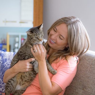 A woman with long hair holds a tabby cat while sitting on a couch.
