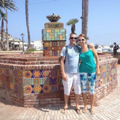 A couple standing together in front of a colorful tiled fountain in a coastal area.