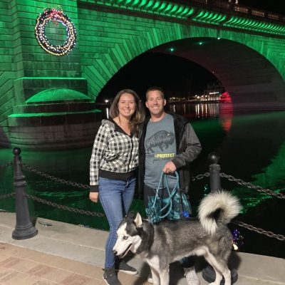 A couple standing with a Siberian Husky dog near a bridge decorated with lights.