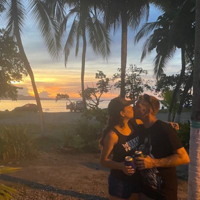 A couple sharing a moment at sunset while holding drinks in a tropical setting.