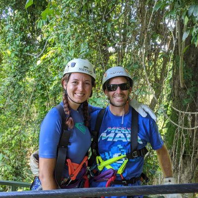 Two individuals wearing zipline harnesses and helmets, smiling at the camera in a forested area.