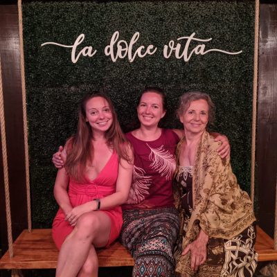 Three women sitting together on a wooden bench in front of a green backdrop with the text 'la dolce vita'.