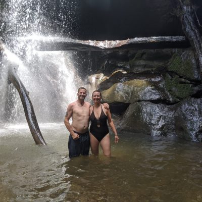 A man and woman standing in shallow water beneath a waterfall in a rocky setting.