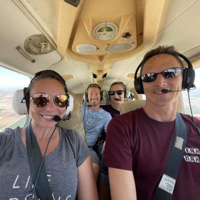 Four individuals seated inside a small aircraft wearing headsets and smiling at the camera.