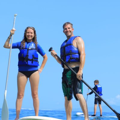 Two individuals wearing life jackets paddleboarding in the ocean on a clear day.
